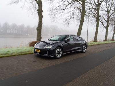 Rijdende Hyundai IONIQ 6 met bomen en een rivier langs de weg.