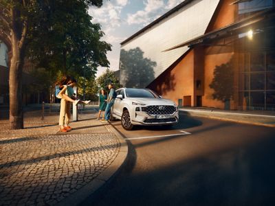 Nowy 7-miejscowy SUV Hyundai SANTA FE Plug-in Hybrid zaparkowany na miejskiej ulicy i opierający się o niego ludzie.