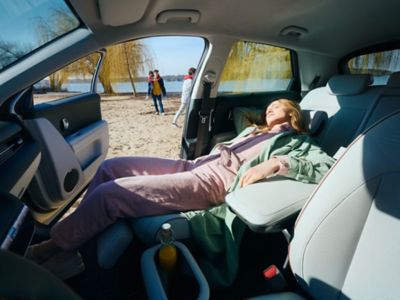 Eine Frau liegt schlafend auf dem zurückgeklappten Relax-Sitz eines an einem Strand parkenden Hyundai IONIQ 5.