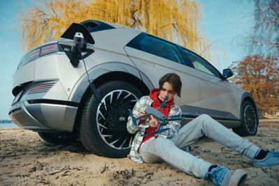 The Hyundai IONIQ 5 electric CUV parked on the beach, with a teenager charging their drone from the car.