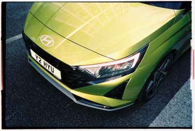 The front of a green Hyundai i20 seen from above driving on a road.	