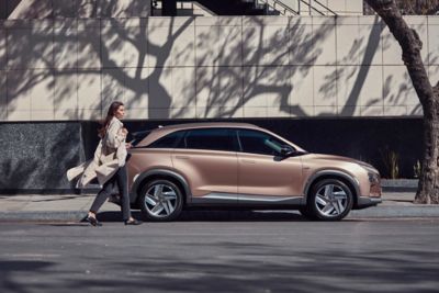 Eine Frau mit wehendem Mantel geht auf einen am Straßenrand geparkten Hyundai NEXO zu.