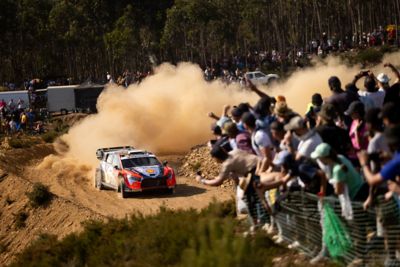 Der Hyundai i20 N Rally1 fährt bei der Portugal-Ralley vor jubelnden Fans in eine Kurve und zieht eine Staubwolke hinter sich her. 