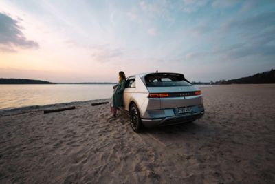 IONIQ 5 zaparkovaný na pláži, o ktorý sa opiera žena počas západu slnka.