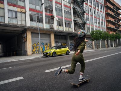 A girl riding a skateboard on the street next to the Hyundai i20 in lucid green. 