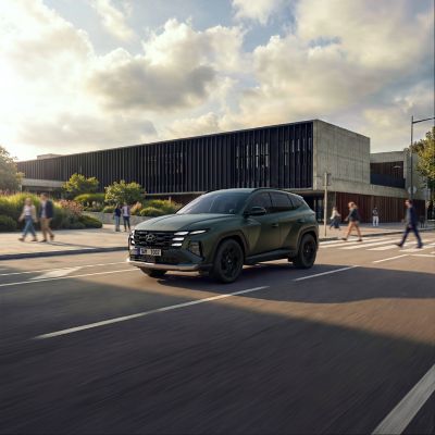 Čelní pohled na Hyundai TUCSON jedoucí po ulici s moderní budovou a chodci v pozadí.