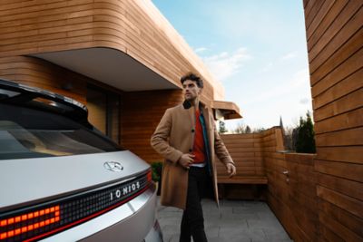 Mann geht zu seinem Hyundai IONIQ 5 in der Einfahrt eines modernen, holzverkleideten Hauses.