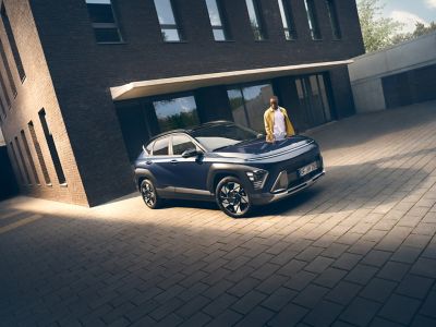 A man leaning on the front of the Hyundai KONA Electric with a city skyline in the back.