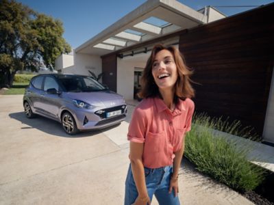 Ein Hyundai i10 parkt in der Einfahrt eines modernen Wohnhauses. Im Vordergrund lacht eine Frau in roter Bluse und Jeans.