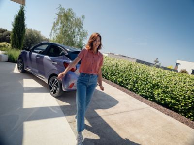 Ein parkender, blauer Hyundai i10 in einer Einfahrt und eine Frau davorstehend.