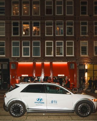 Zijaanzicht witte Hyundai IONIQ 5 met Hyundai-logo en Amsterdam Fashion Week-logo op portieren, met op achtergrond een Amsterdams gebouw.