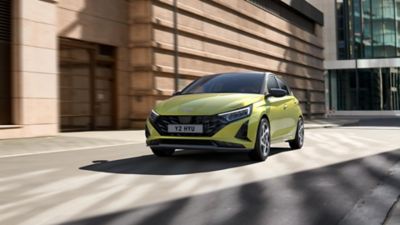 A green Hyundai i20 driving between modern building blocks.	