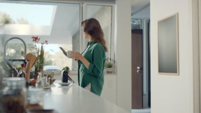 En kvinne står på kjøkkenet med smarttelefonen i en hånd og en kopp i den andre. En Hyundai står i oppkjørselen. Foto.