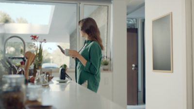 A woman at a kitchen counter, mug in one hand, smartphone in the other, a Hyundai parked in her driveway.	