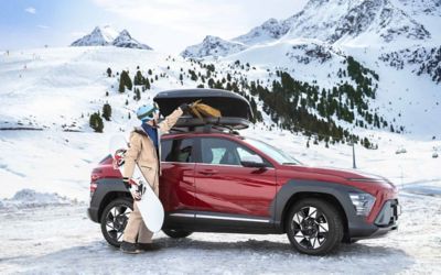 Man in wintersportoutfit met snowboard in de hand naast een rode Hyundai KONA op een besneeuwde berg