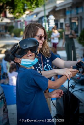 Ein Junge mit einer VR-Brille und eine Frau mit Sonnenbrille, die ihm bei der Bedienung hilft.