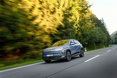 Driekwart vooraanzicht Hyundai KONA Electric rijdend op een weg in bosrijke omgeving.