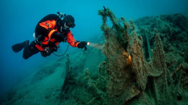 Buceador bajo el agua con una linterna, inspeccionando el lecho marino en busca de redes de pesca