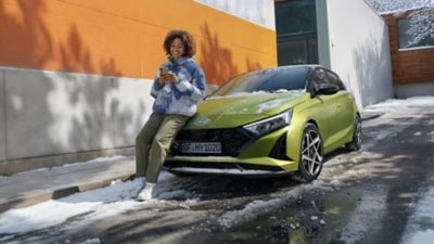 Eine Frau am Handy lehnt an der Motorhaube eines parkenden i20. 