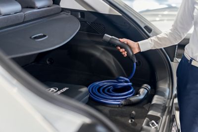 Ein Mode-3-Ladekabel liegt im geöffenten Kofferraum eines Hyundai Fahrzeugs, ein Steckerende wird hoch gehalten.