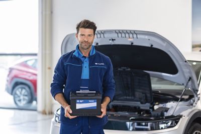 Ein Hyundai Service-Techniker steht mit einer Autobatterie in den Händen vor einem Hyundai Fahrzeug mit geöffneter Motorhaube.