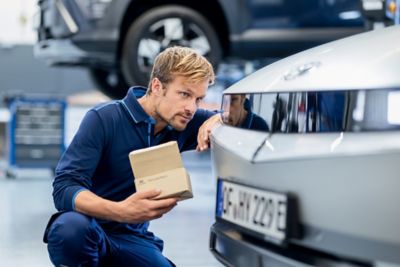 Ein Hyundai Service-Techniker mit einem kleinen Karton in der Hand wirft einen prüfenden Blick auf das Vorderlicht eines Hyundai Fahrzeugs.