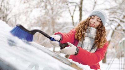 Eine Frau im Winteroutfit befreit die Scheibe eines Hyundai Fahrzeugs von Eis und Schnee.