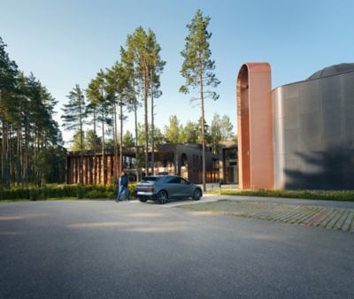 Ein Hyundai IONIQ 5  steht an einer Ladestation in der Einfahrt moderner Häuser. Der Fahrer geht lachend und entspannt am Fahrzeug vorbei, während warmes Sonnenlicht durch die hohen Bäume fällt.
