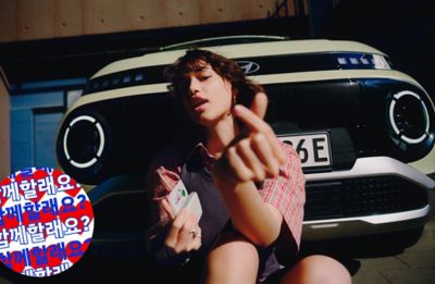 Eine junge Frau sitzt vor dem Hyundai INSTER auf dem Boden und zeigt mit der linken Hand ein Herz-Symbol. Links unten im Bild befindet sich ein runder Sticker mit koreanischen Schriftzeichen.