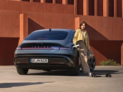 Eine Frau lehnt sich an einen IONIQ 6 und lädt ein elektrisches Skateboard, das an den externen Ladeanschluss angeschlossen ist.