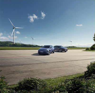 Zwei Hyundai KONA, schräg von vorne gesehen, parken auf einer Freifläche vor einer grünen Landschaft mit Windrädern und Bergkulisse.