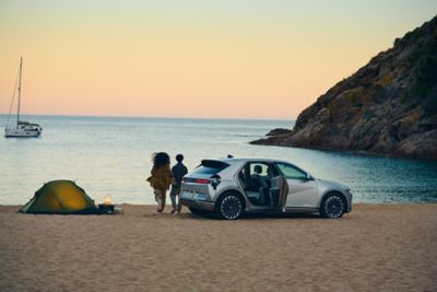 Ein Paar mit ihrem IONIQ 5 am Strand bei Sonnenuntergang. Neben ihnen steht ein Zelt.