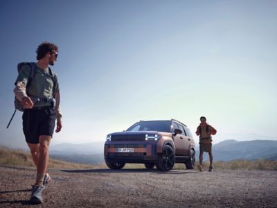 Ein Wanderer steht auf einem Bergpfad neben einem Hyundai SANTA FE mit Blick auf eine weite, bergige Landschaft.