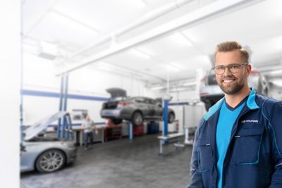 Ein Hyundai-Service-Techniker steht lächelnd in einer Autowerkstatt,.im Hintergrund befindet sich ein Auto auf einer Hebebühne.