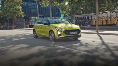 Ein Hyundai i20 in der aufpreispflichtigen Farbe Lucid Lime Metallic fährt auf einer von Bäumen beschatteten Straße.