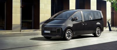 Der Hyundai STARIA, schräg von vorne gesehen,  steht vor einem innerstädtischen Bürogebäude. 