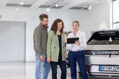 Eine Hyundai Mitarbeiterin berät in einem Autohaus mit Hilfe eines Tablets ein Paar, daneben steht ein IONIQ 5.