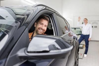 Ein Mann fährt nach einem Hyundai Servicetermin lächelnd aus der Werkstatt. Im Hintergrund steht eine Hyundai Servicemitarbeiterin.