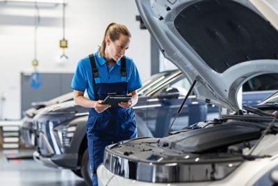 Eine Hyundai Servicemitarbeiterin mit Tablet-Computer wirft einen prüfenden Blick in den Motorraum eines Hyundai.