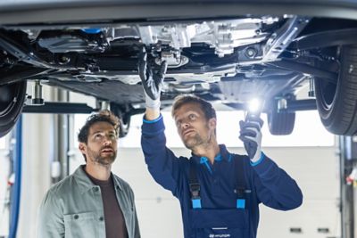 Ein Hyundai Service-Mitarbeiter und ein Kunde stehen unter einer Hebebühne und sehen sich den Unterboden eines Hyundai Fahrzeugs an.