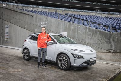 Eine Frau im roten Eintracht Frankfurt Trikot steht an der Beifahrerseite eines Hyundai KONA Elektro, der neben der Stadiontribüne steht.