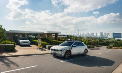 Ein Hyundai Robotaxi fährt an einem Wohnhaus vorbei, in dessen Einfahrt ein Hyundai IONIQ 5 und eine Frau mit dem Roboterhund Spot zu sehen sind.