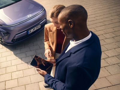 Ein Mann und eine Frau stehen vor einem Hyundai Fahrzeug und schauen auf ein Tablet.