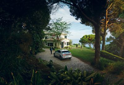 Ein Hyundai IONIQ 5 auf der Zufahrt zu einem Haus an einer Steilküste, in dessen Garten eine Familie mit einem Hund spielt. 