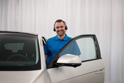 Eine Online-Showroom-Mitarbeiterin mit Headset steht neben einem Hyundai NEXO und erklärt seine Vorzüge.