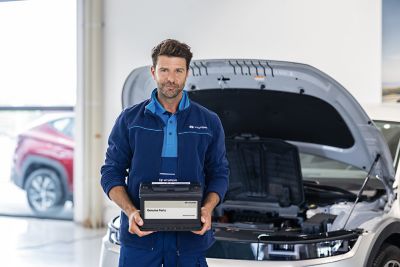 Hyundai-tekniker håller ett Hyundai originalstartbatteri framför ett öppet motorutrymme.