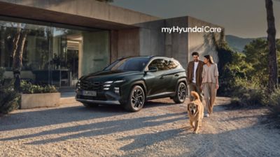 Mörk Hyundai TUCSON Plug-in Hybrid parkerad framför modernt hus och ett ungt par med hund som går bredvid.