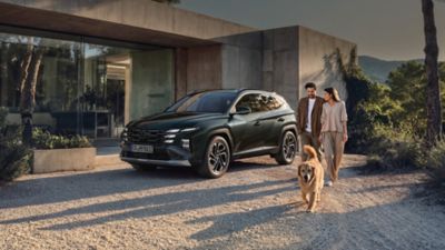 Mörk Hyundai TUCSON Plug-in Hybrid parkerad framför modernt hus och ett ungt par med hund som går bredvid.