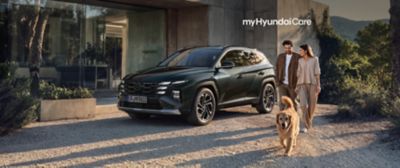 Mörk Hyundai TUCSON Plug-in Hybrid parkerad framför modernt hus och ett ungt par med hund som går bredvid. 