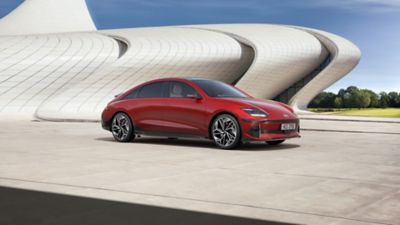Driekwart vooraanzicht Hyundai IONIQ 6 in de opvallende kleur rood, geparkeerd voor een futuristisch gebouw.